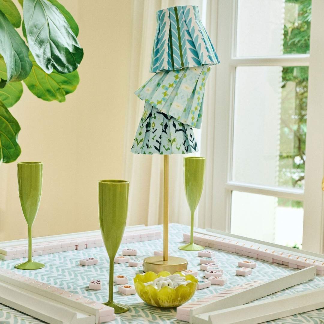 Chloe Blue box pleat lamp shade cover by Laura Park with a hand-painted floral pattern in shades of blue, cream, and olive with a green and white patterned shade on a mahjong table with green champagne flutes and a plant.
