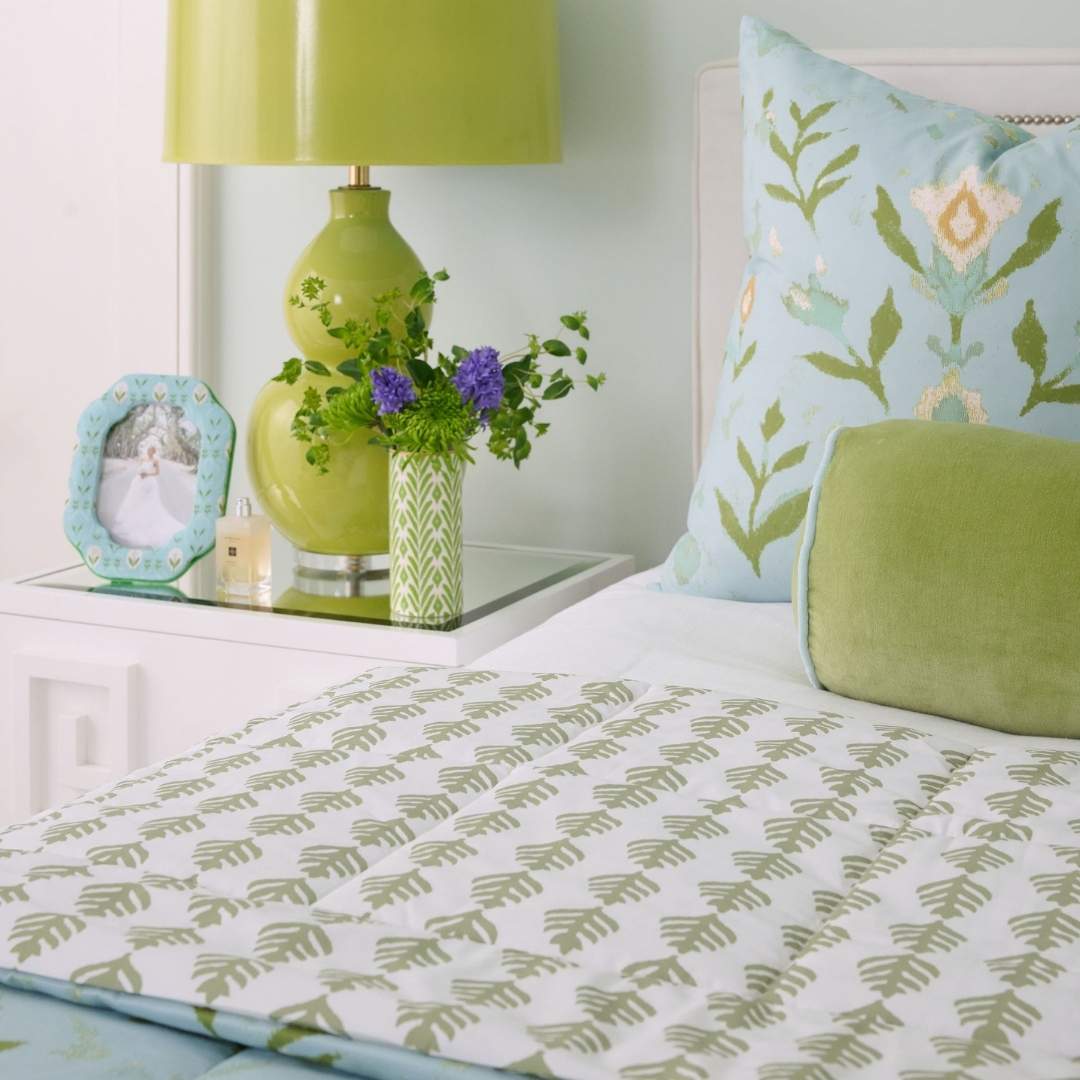 blue, green, and white hand painted floral and leaf patterned bedding set designed by Laura Park pillows, lamp, frame and vase in the background, styled for a guest room, bedroom, or dorm room.