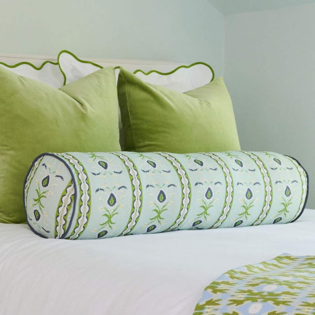 Laura park rosemary stripe decorative cylindrical bolster pillow with green and white floral hand painted pattern seen on a white bed with scalloped green shams, velvet pillows, and a green and blue willow weave throw pillow.