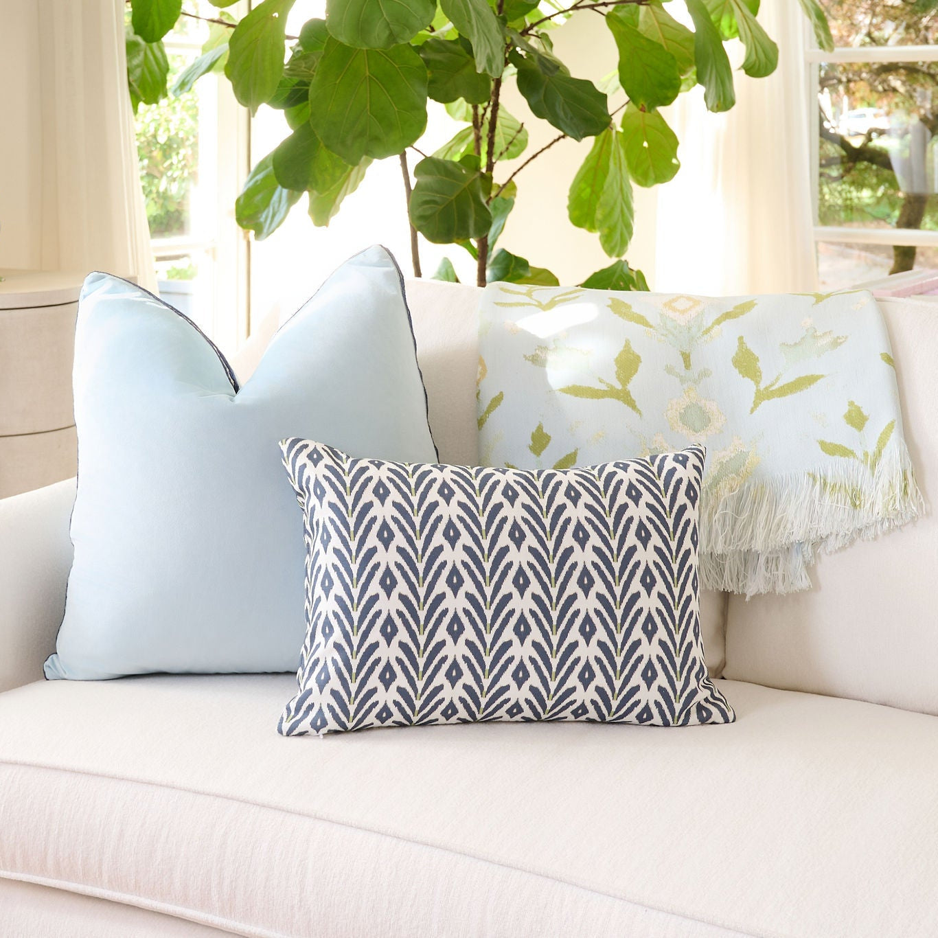 Laura Park Petal Garden Aqua Throw Blanket styled with a petite frond navy pillow and blue velvet pillow on a white beige couch in a well lit living room.