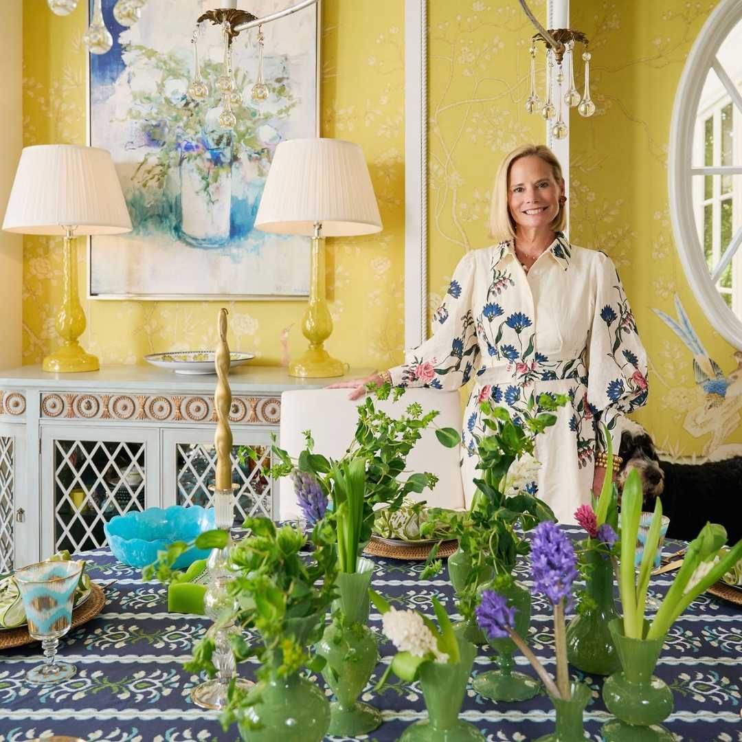 A woman stands in a bright yellow room behind a decorated table with green glass vases, flowers, lamps, and a blue patterned tablecloth. She is wearing a cream floral dress, and the room has yellow wallpaper and framed artwork.