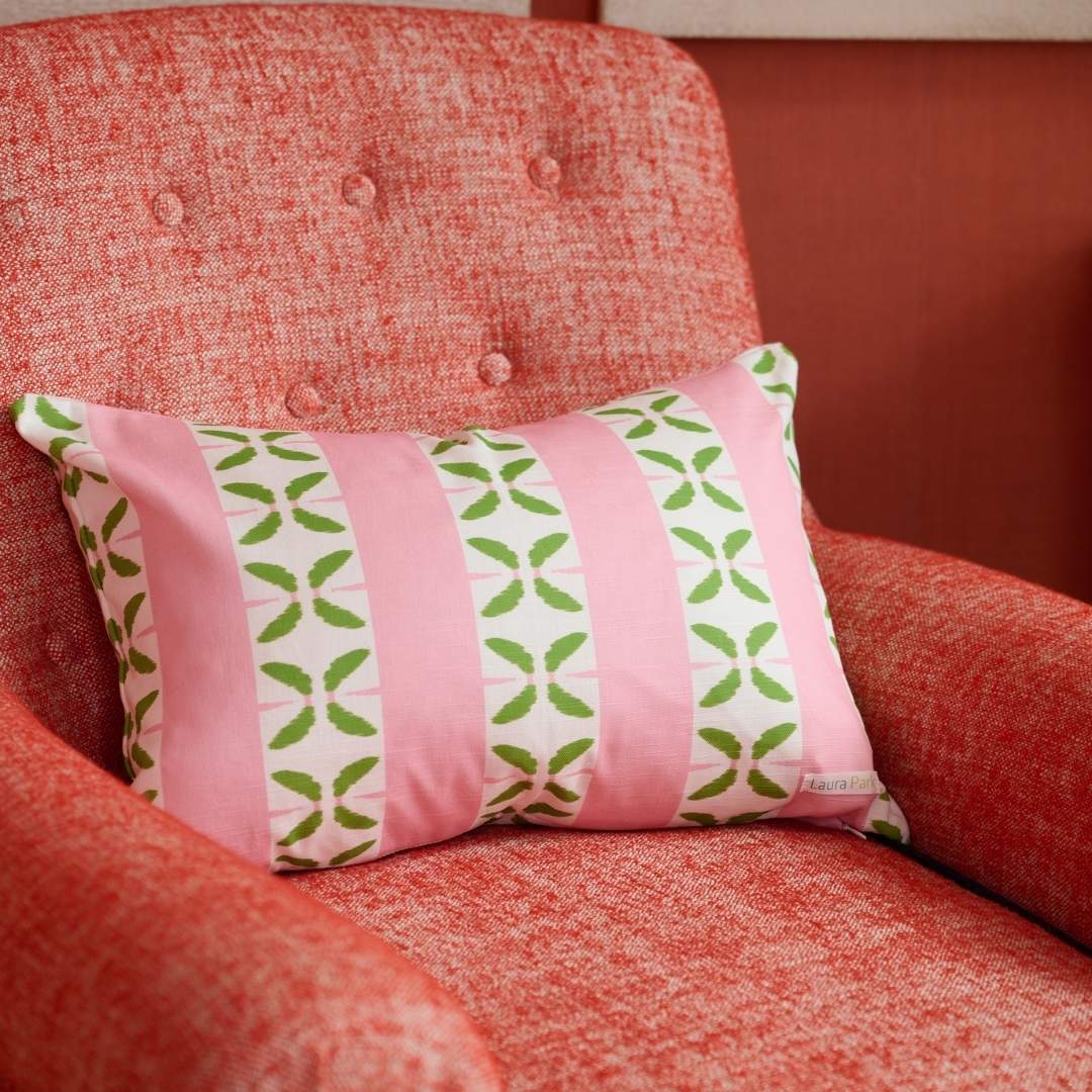 Laura Park Petal Stripe Pink decorative throw pillow with pink and white stripes and green leaf patterns styled on a red accent reading chair in the corner of a room with red walls and white frames in the background. 