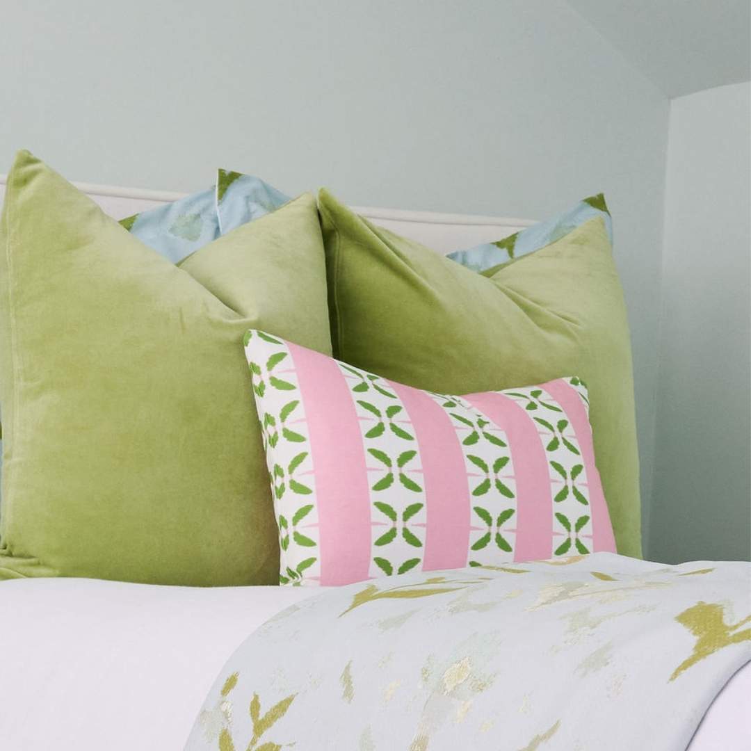 Laura Park Petal Stripe Pink decorative throw pillow with pink and white stripes and green leaf patterns styled on a white bed with green velvet pillows and petal garden aqua shams and duvet cover in a room with light blue walls. 