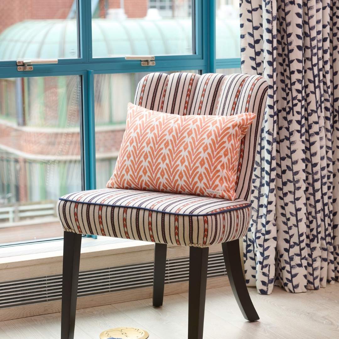 Decorative pillow with ikat inspired orange and white geometric painted pattern by Laura Park Designs styled on a striped scalloped accent chair in front of a blue paned window with navy and white curtains. 