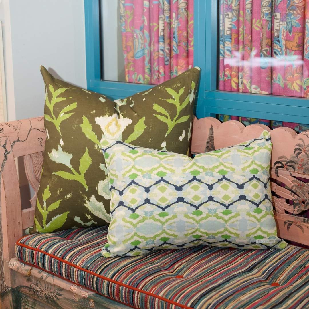 Petal Garden Olive decorative throw pillow by Laura Park Designs featuring hand-painted green and blue florals in light blue and green on a deep olive ground paired with Laura Park's Willow Weave decorative throw pillow on a pink indoor bench.