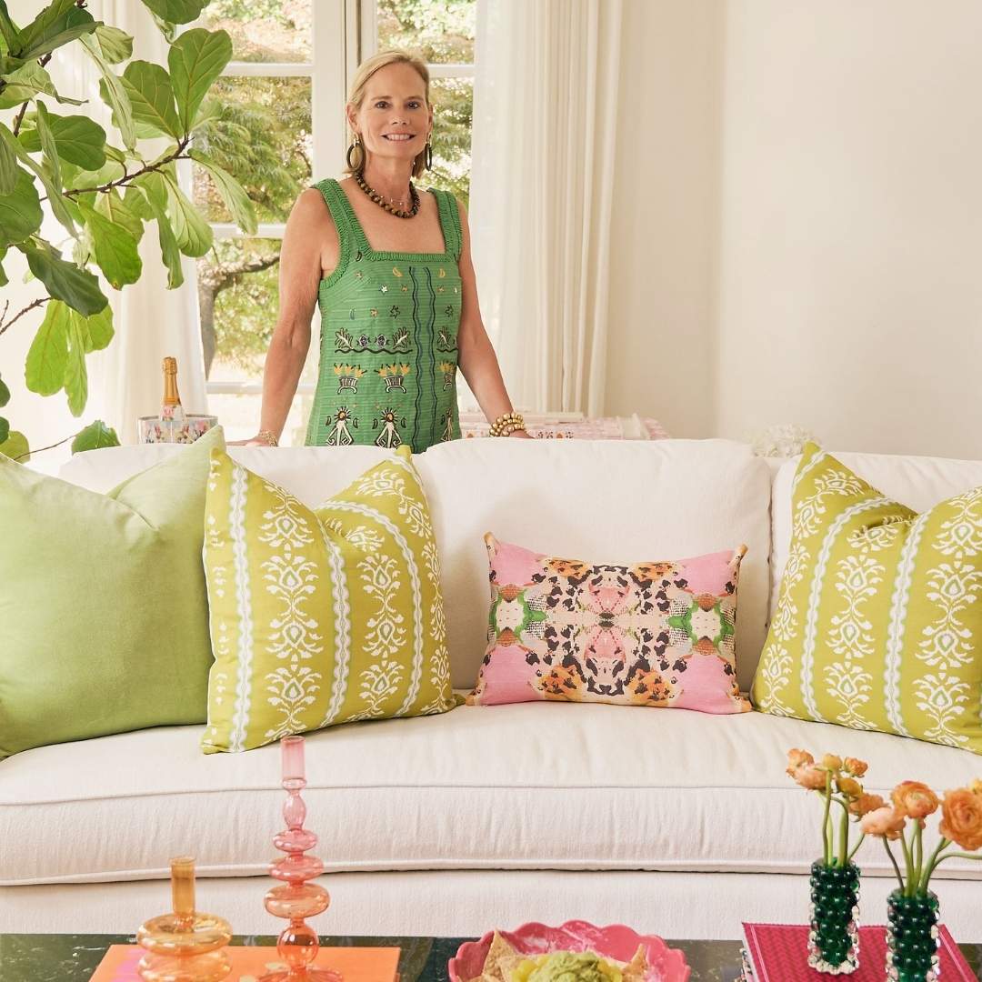 Laura Park standing behind a light colored couch with pillows from her Cotswold Collection launching in June 2026 including the Tiger Lily, Rosemary Stripe, and Green Velvet decorative throw pillows. 
