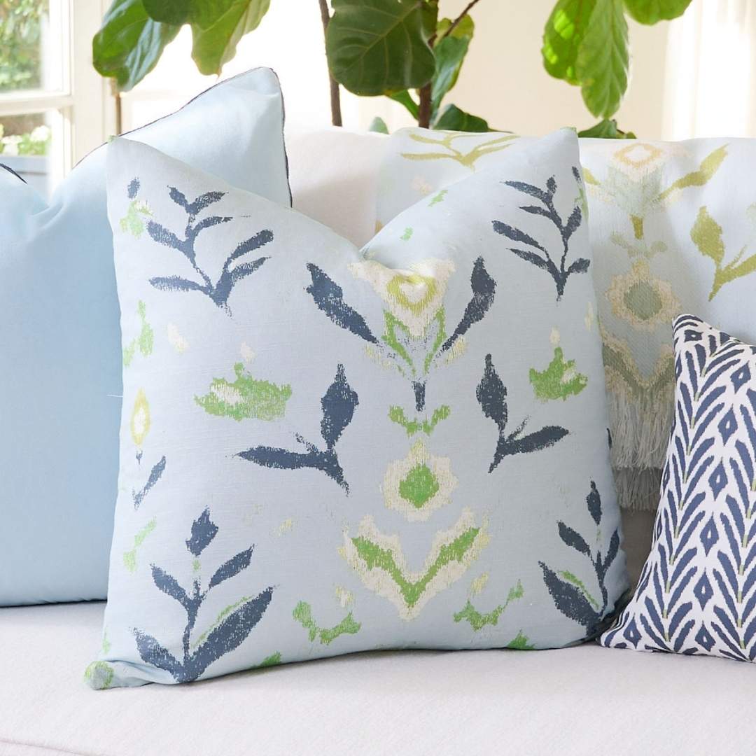 Decorative pillow with a navy, olive green, and off white handpainted floral pattern on a light blue background styled on a grey couch with a blue velvet pillow and a petite frond navy decorative throw pillow. 