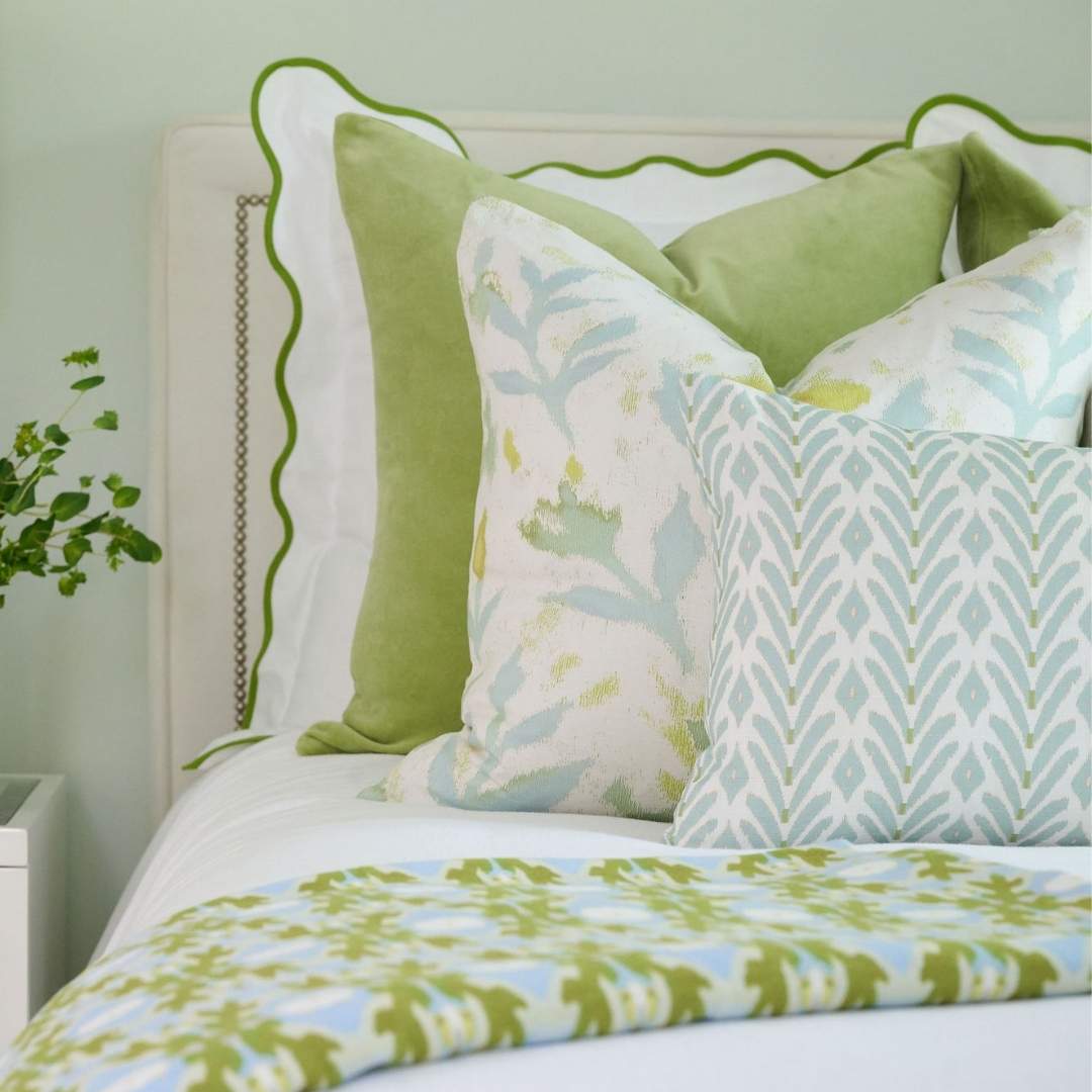 An ikat-inspired motif in soft blue with subtle green accents, Laura Park's Petite Frond Blue Decorative Pillow is styled with the petite garden linen, green velvet, and scalloped white and green euro shams on a white bed with a blue and green.