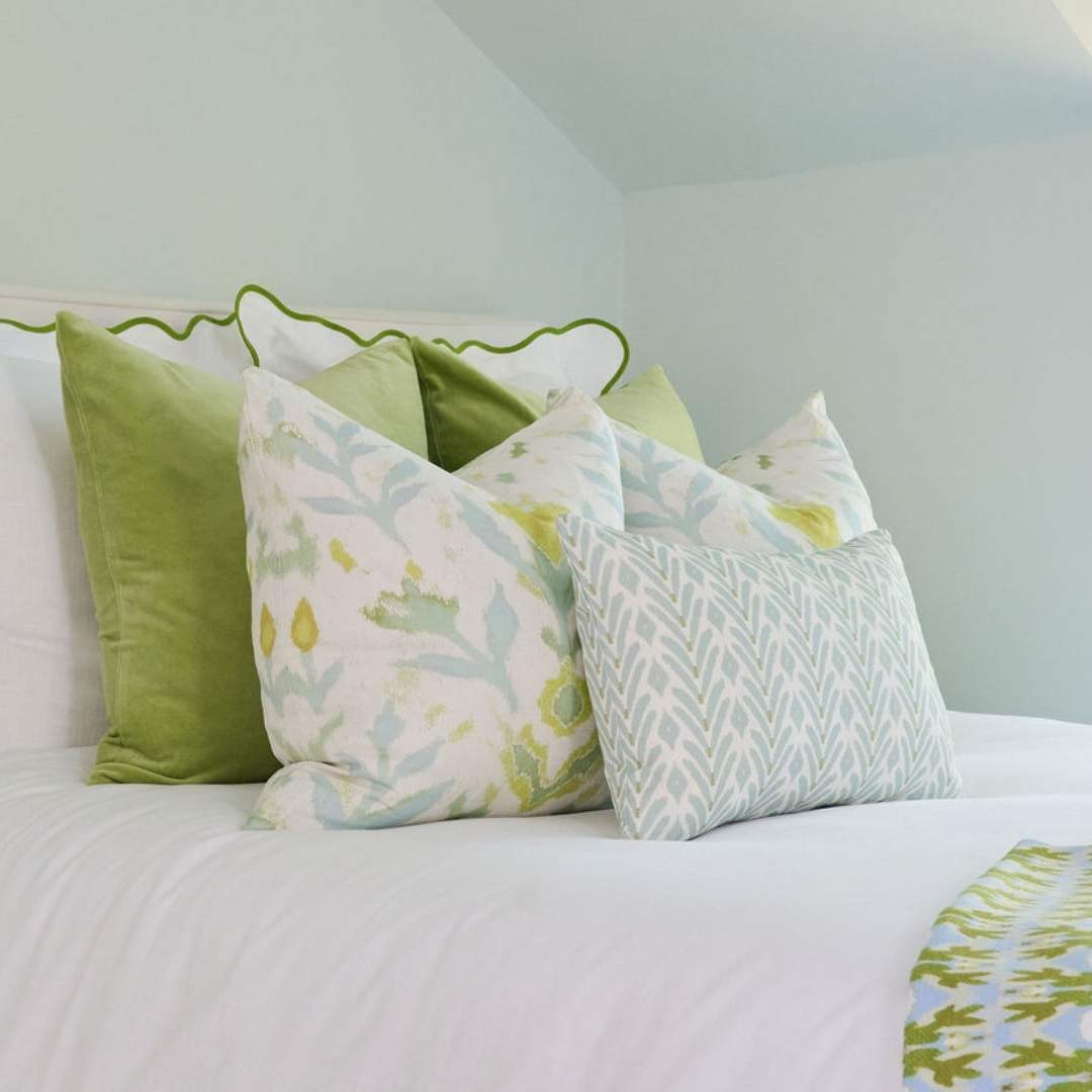 Laura Park's signature painted design, Petal Garden Linen (white pillow with green and blue accents and a floral design), on a decorative pillow styled with a white bedspread with green accents,  velvet throw pillows. the petite frond blue.