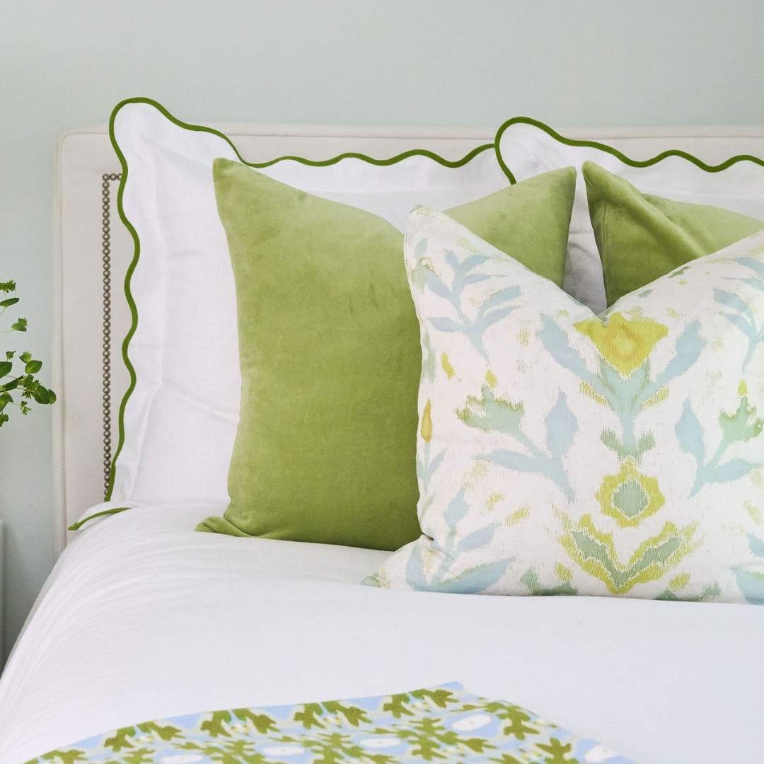 Laura Park's signature painted design, Petal Garden Linen (white pillow with green and blue accents and a floral design), on a decorative pillow styled with a white bedspread with green accents and velvet throw pillows. 