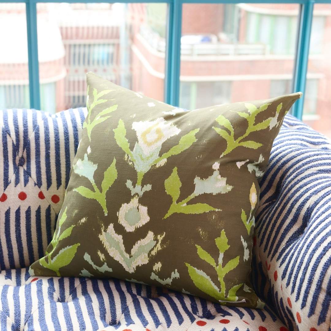 Petal Garden Olive decorative throw pillow by Laura Park Designs featuring hand-painted green and blue florals in light blue and green on a deep olive ground styled on a blue patterned couch in front of a bright blue window. 