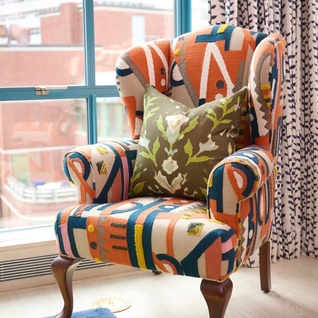 Petal Garden Olive decorative throw pillow by Laura Park Designs featuring hand-painted green and blue florals in light blue and green on a deep olive ground styled on a patterned club accent chair in front of a blue window with patterned navy.