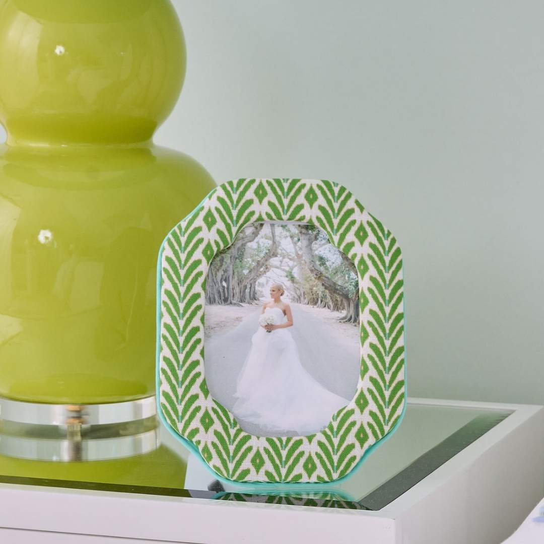 The Petite Frond Fabric Frame with ikat-inspired motif in bright green with subtle blue seen here with a bridal portrait inside the frame styled on a white bedside table next to a green contemporary lamp. 