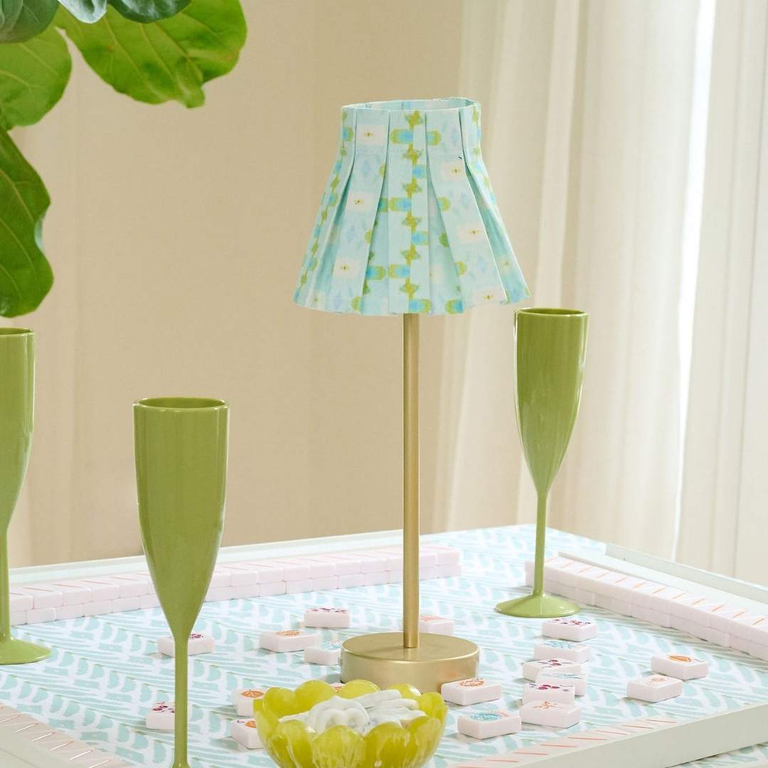 Chloe Blue box pleat lamp shade cover by Laura Park with a hand-painted floral pattern in shades of blue, cream, and olive on a table with green champagne flutes. 