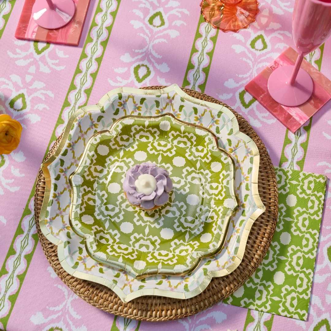 A styled tabletop setting featuring a stack of scalloped paper plates with a green and white damask pattern and gold trim, placed on woven placemats over a pink patterned tablecloth with pink glassware and floral accents.