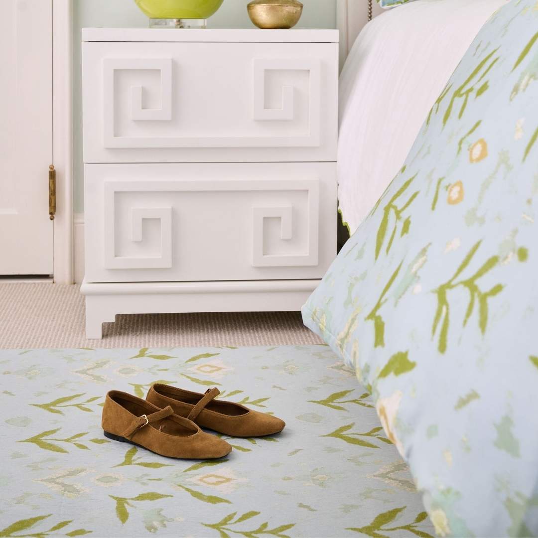 Laura Park's Signature painted pattern, petal garden aqua on a floor mat rug for indoor use in kitchen, bathroom, entry room, mud room, bedroom. Seen here in a bedroom next to a bed and nightstand with brown suede flats on top of the mat. 