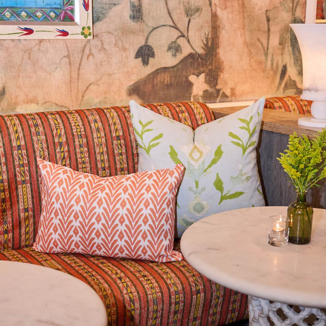 Decorative pillow with ikat inspired orange and white geometric painted pattern by Laura Park Designs on a patterned red and yellow couch paired with the laura park petal garden aqua throw pillow. 
