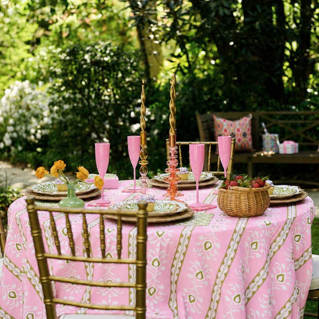 A round outdoor table set with a pink patterned tablecloth, pink champagne flutes, green plates, and gold-toned chairs, surrounded by lush green garden foliage.
