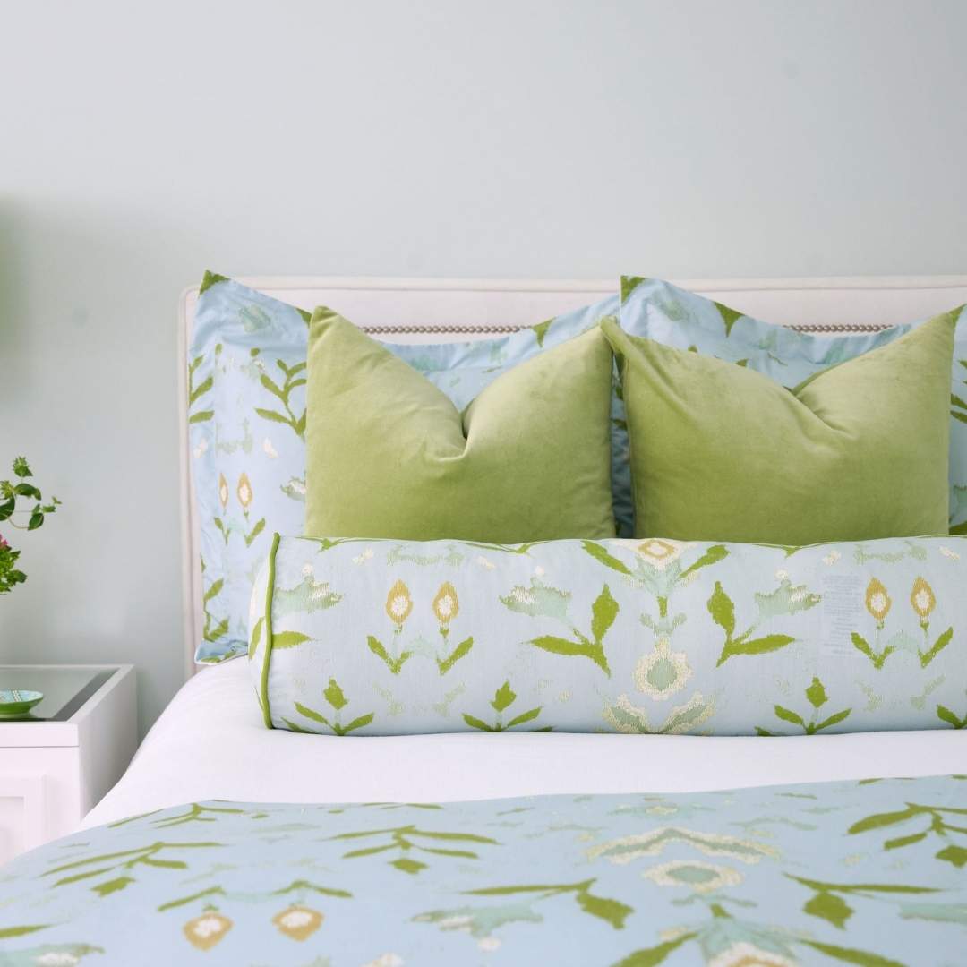 Round bolster pillow with Laura Park's Petal Garden Aqua, a hand painted design with blue, olive, and cream motifs with a green embroidered border styled on a white bed with green velvet pillows and petal garden shams in a blue and green.
