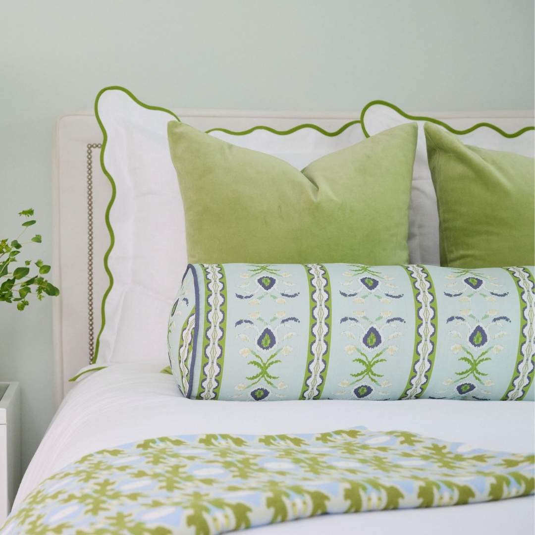 Laura park rosemary stripe decorative cylindrical bolster pillow with green and white floral hand painted pattern seen on a white bed with scalloped green shams, velvet pillows, and a green and blue willow weave throw pillow. 