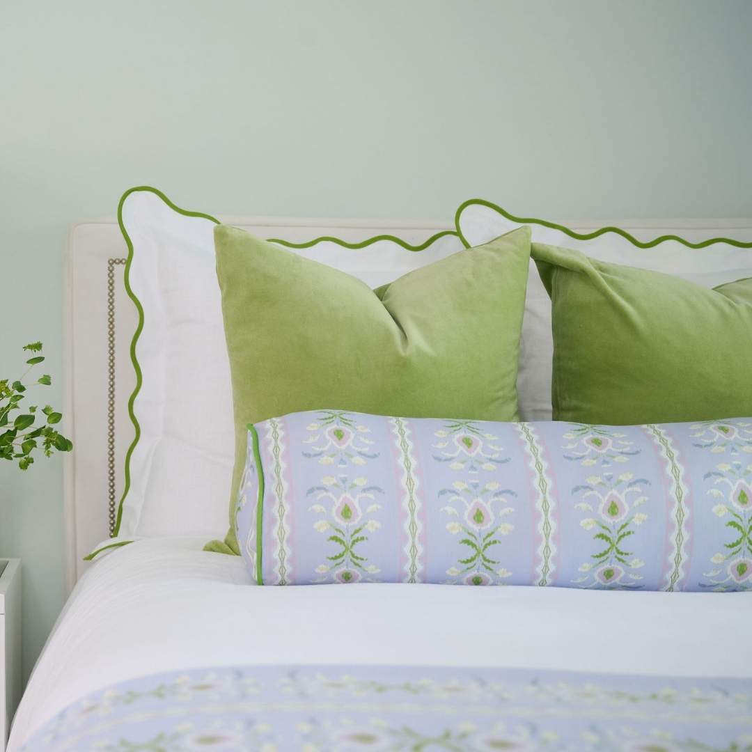 Decorative cylindrical laura park bolster pillow with floral pattern on a light blue lavender background and green piping styled with velvet green pillows and green scalloped euro shams. 