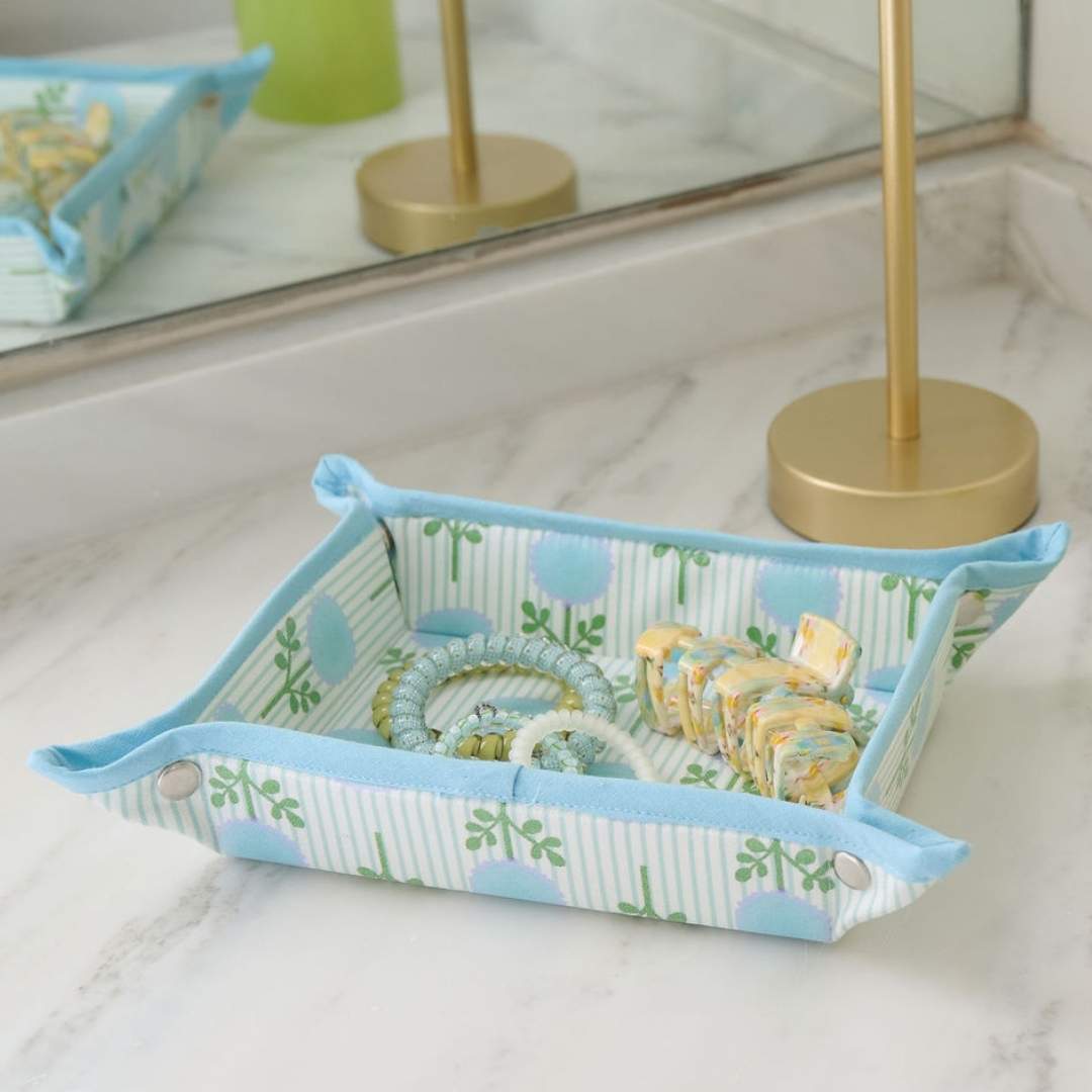 Decorative tray holding jewelry and hair accessories with a floral pattern on a marble surface. Pattern contains blue hand painted marigolds overlayed over light blue ticking stripes. 