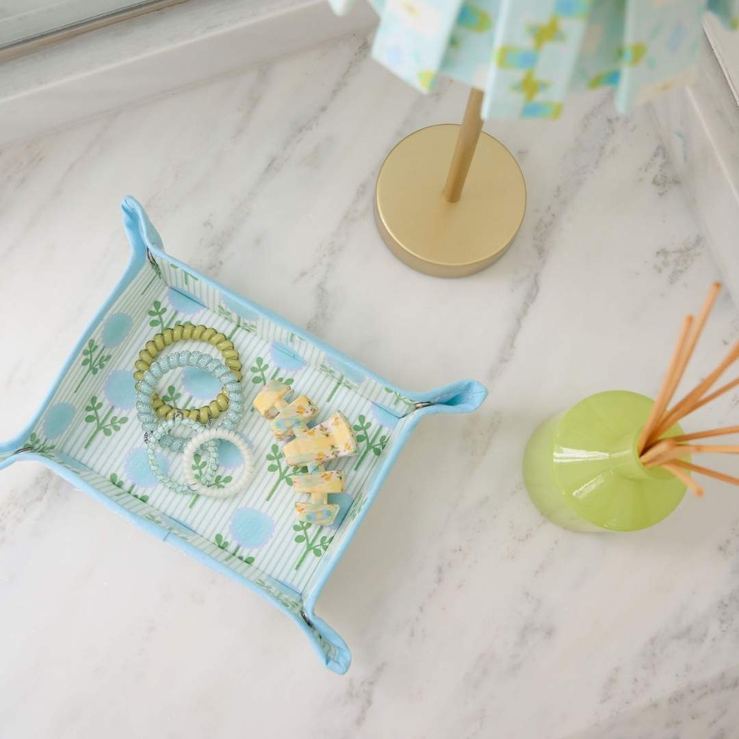 Decorative tray holding jewelry and hair accessories with a floral pattern on a marble surface. Pattern contains blue hand painted marigolds overlayed over light blue ticking stripes. 