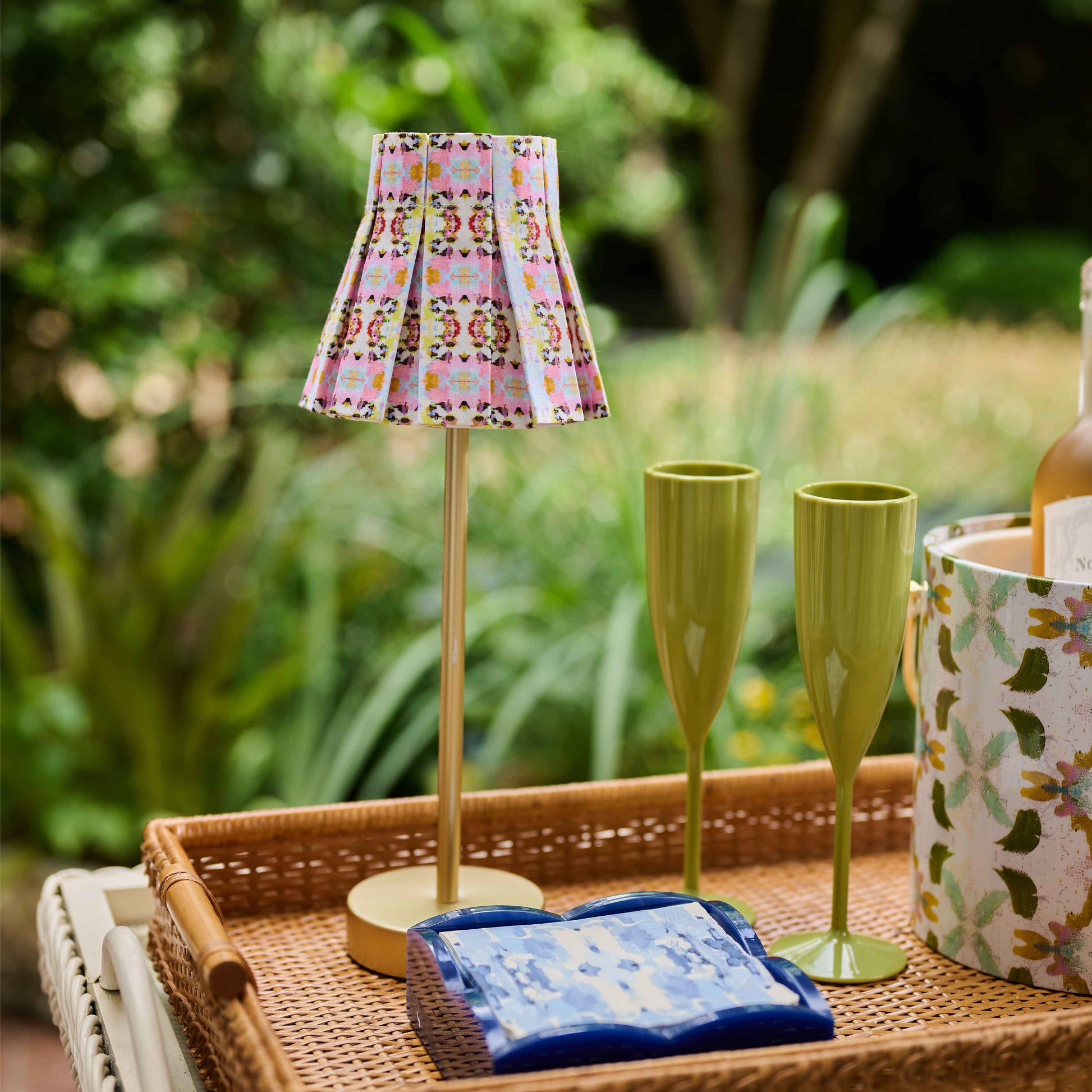 A styled outdoor patio scene featuring a woven wicker tray on a white wicker table. The tray contains a miniature table lamp with a pink and yellow patterned shade, two olive green fluted glasses, a blue patterned napkin, and a floral-patterned bucket bucket.