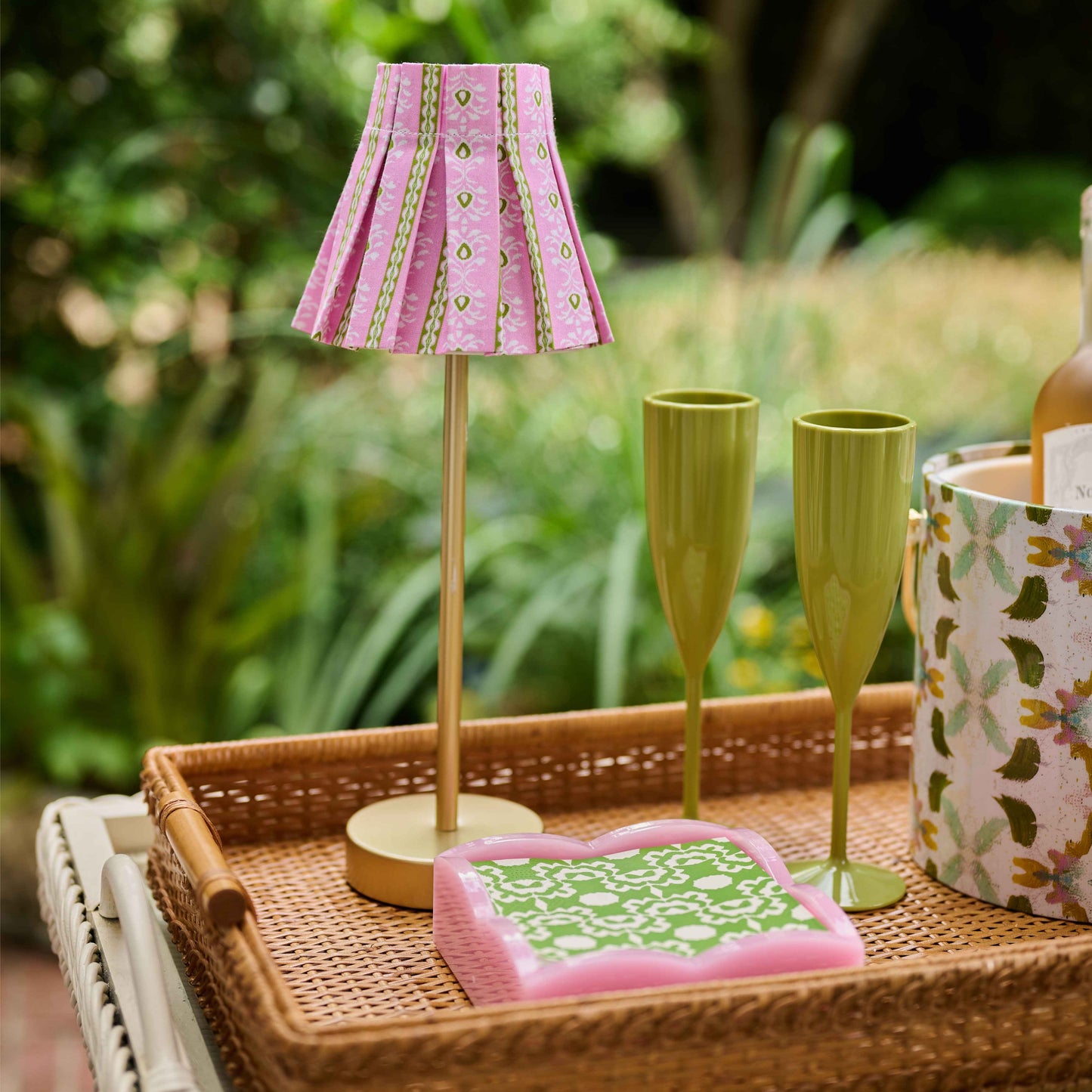 A styled tabletop scene with a woven tray holding a small pink patterned table lamp, two green fluted glasses, a pink and green patterned book or notebook, and a floral-patterned container, set against a blurred green garden background.