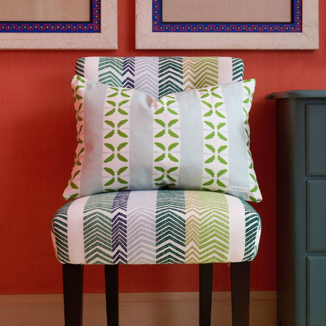 Laura Park's Petal Stripe Light Blue Decorative Throw Pillow with blue stripes and painterly green petal motifs rstyled here on a striped chevron patterned accent chair next to a dark teal side table in front of a red wall. 